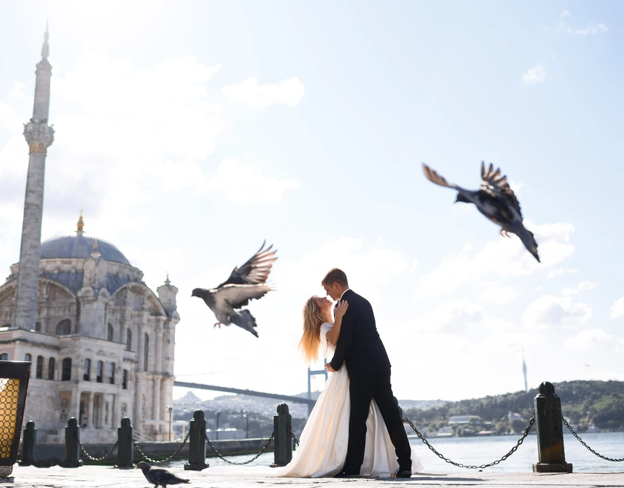 Side view of loving couple posing on quay near Ortakoy mosque in Istanbul while doves flying above, attractive groom in black suit holding bride in wedding dress and looking