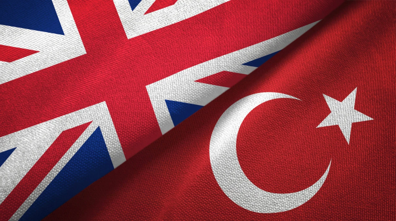 Image showing the flags of the United Kingdom and Türkiye displayed next to each other on a textured fabric background. The UK flag (Union Jack) appears on the left with its distinctive red, white, and blue design, while the Turkish flag on the right features a white crescent moon and star on a red background