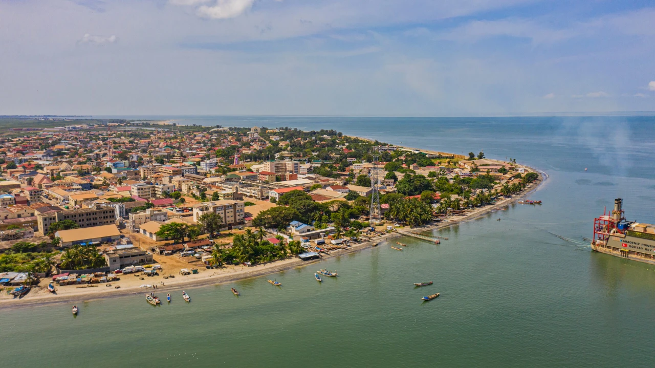 The aerial view shows Banjul, the capital city of The Gambia, with its coastal port and harbor facilities. The image captures the city's layout with colorful buildings extending to the coastline, where several small fishing boats are visible in the water. A larger port structure appears on the right side of the frame. The peninsula-like geography of Banjul is evident, with water surrounding much of the urban area. Palm trees line portions of the shoreline, and the coastal development shows a mix of residential and commercial structures typical of this West African capital city.