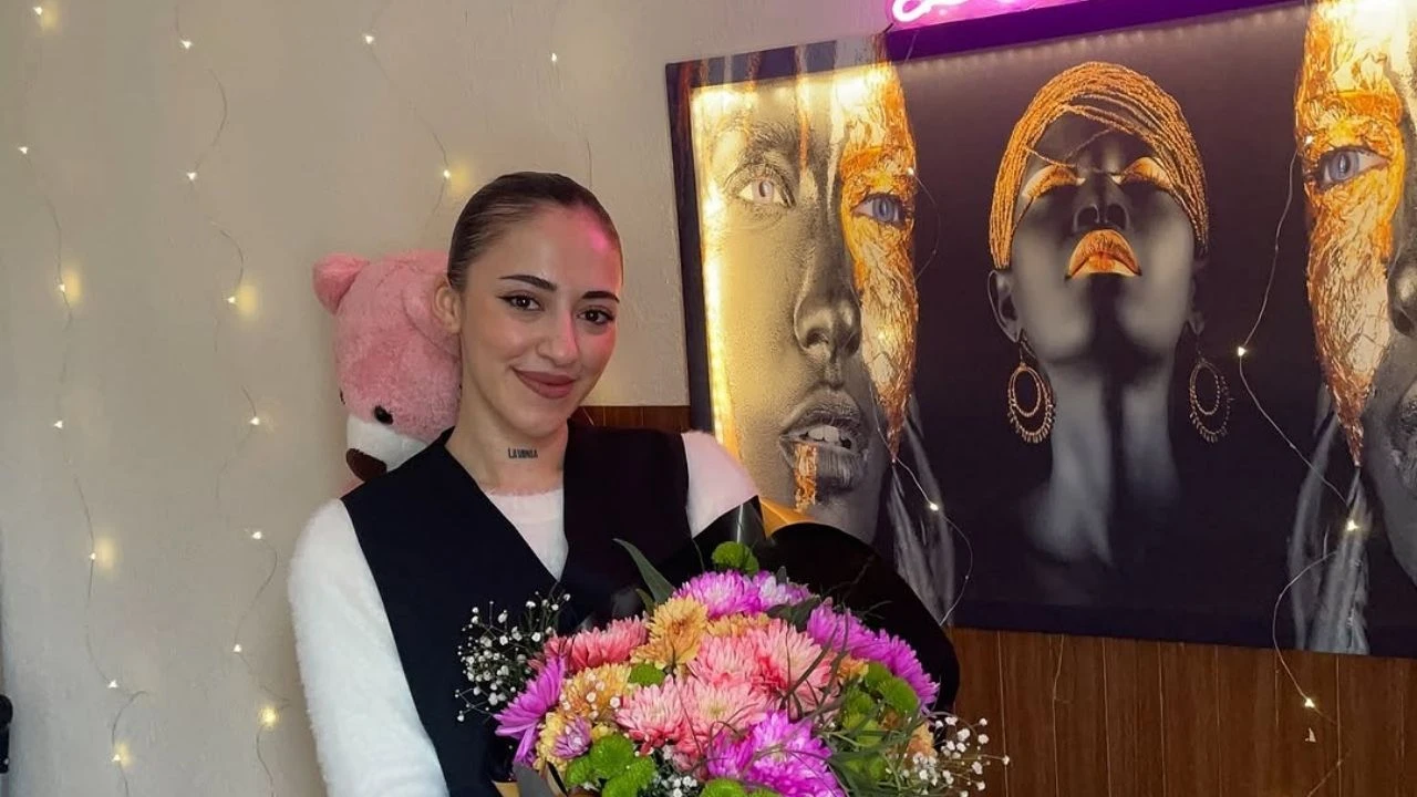 A young woman with a high ponytail smiling while holding a bouquet of flowers in a decorated room with a neon sign and an artistic portrait in the background.