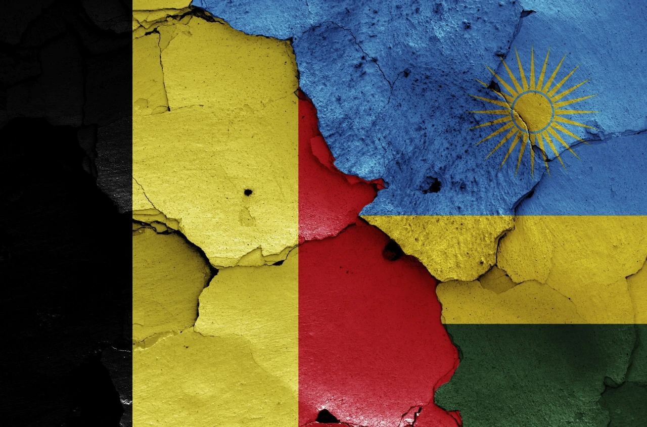 Image shows a textured, cracked wall painted with the national flags of Rwanda (blue, yellow, and green with a sun symbol) and Belgium (black, yellow, and red vertical stripes). The flags appear damaged and fragmented against each other, visually representing the diplomatic rupture between the two countries.