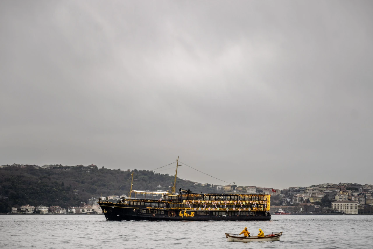 A small wooden boat with two fishermen in yellow raincoats rows in the foreground, while the cityscape of Istanbul