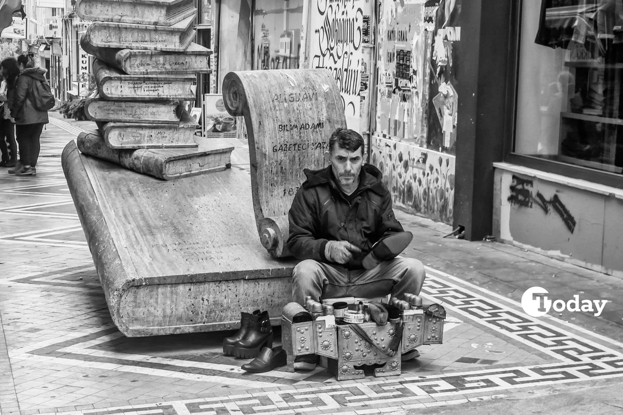 A middle-aged shoe shiner sits on a sidewalk in Kadikoy, Istanbul, with his shoe polishing kit, surrounded by the hustle and bustle of city life. Retirement in Türkiye