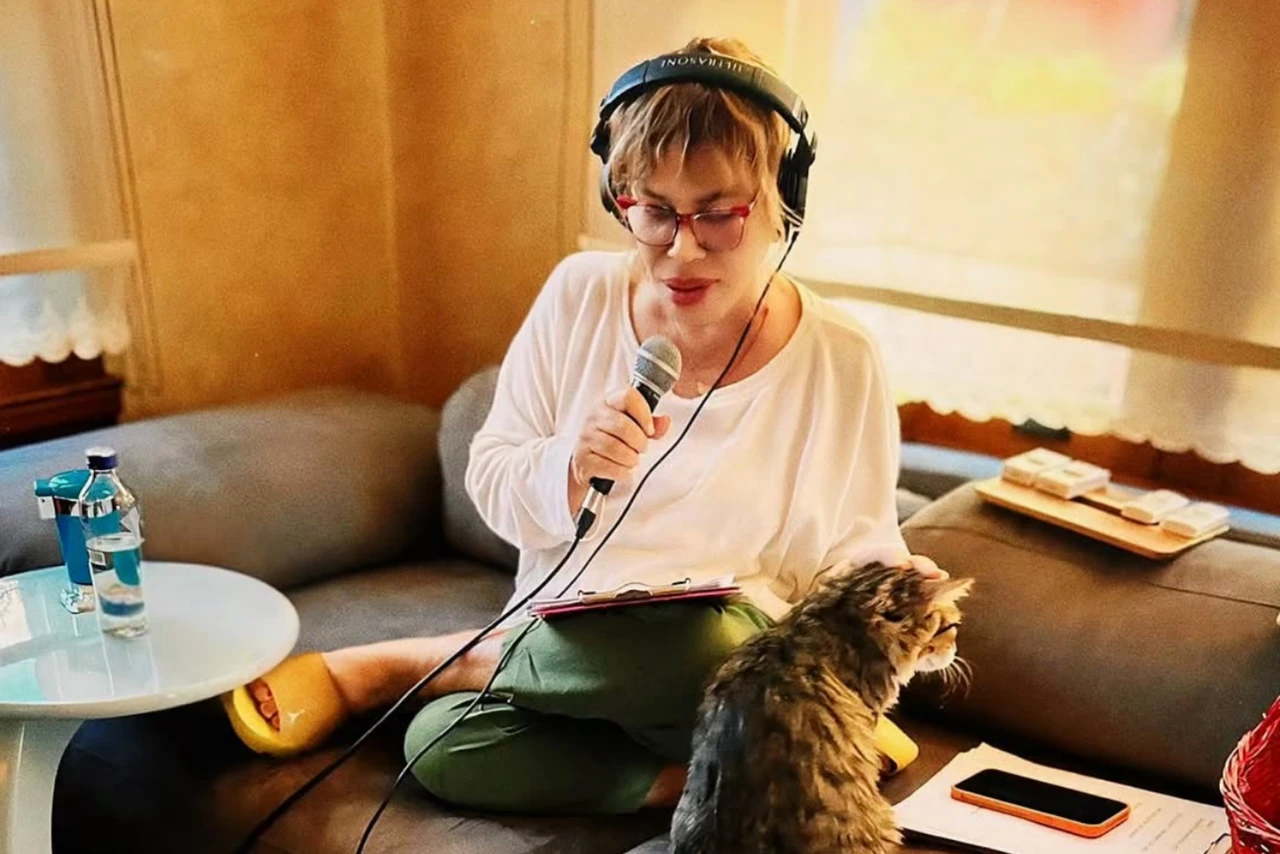 Sezen Aksu sitting on a couch with a microphone and headphones, singing while her cat sits nearby.