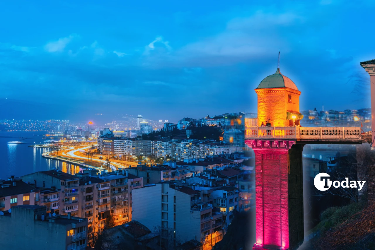 A nighttime view of Izmir, Türkiye, featuring the brightly lit Asansör (Historical Elevator) with its red and orange lights. The cityscape in the background showcases high-rise buildings, winding coastal roads, and reflections of streetlights along the Aegean Sea.