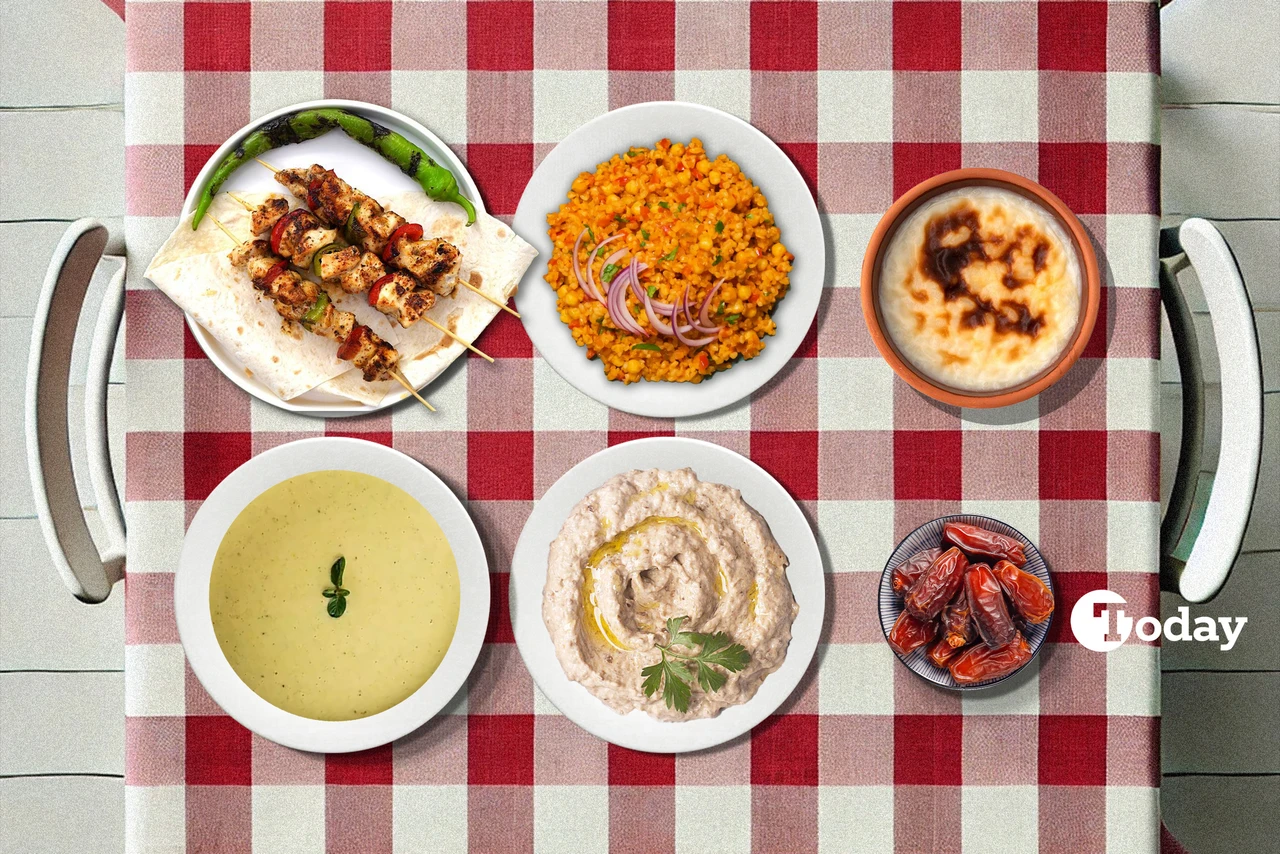 A Turkish iftar meal set on a red and white checkered tablecloth, featuring a bowl of creamy zucchini soup, grilled chicken skewers with flatbread and peppers, a plate of bulgur pilaf garnished with red onions, a smoky eggplant and yogurt dip, oven-baked rice pudding, and a small bowl of dates.