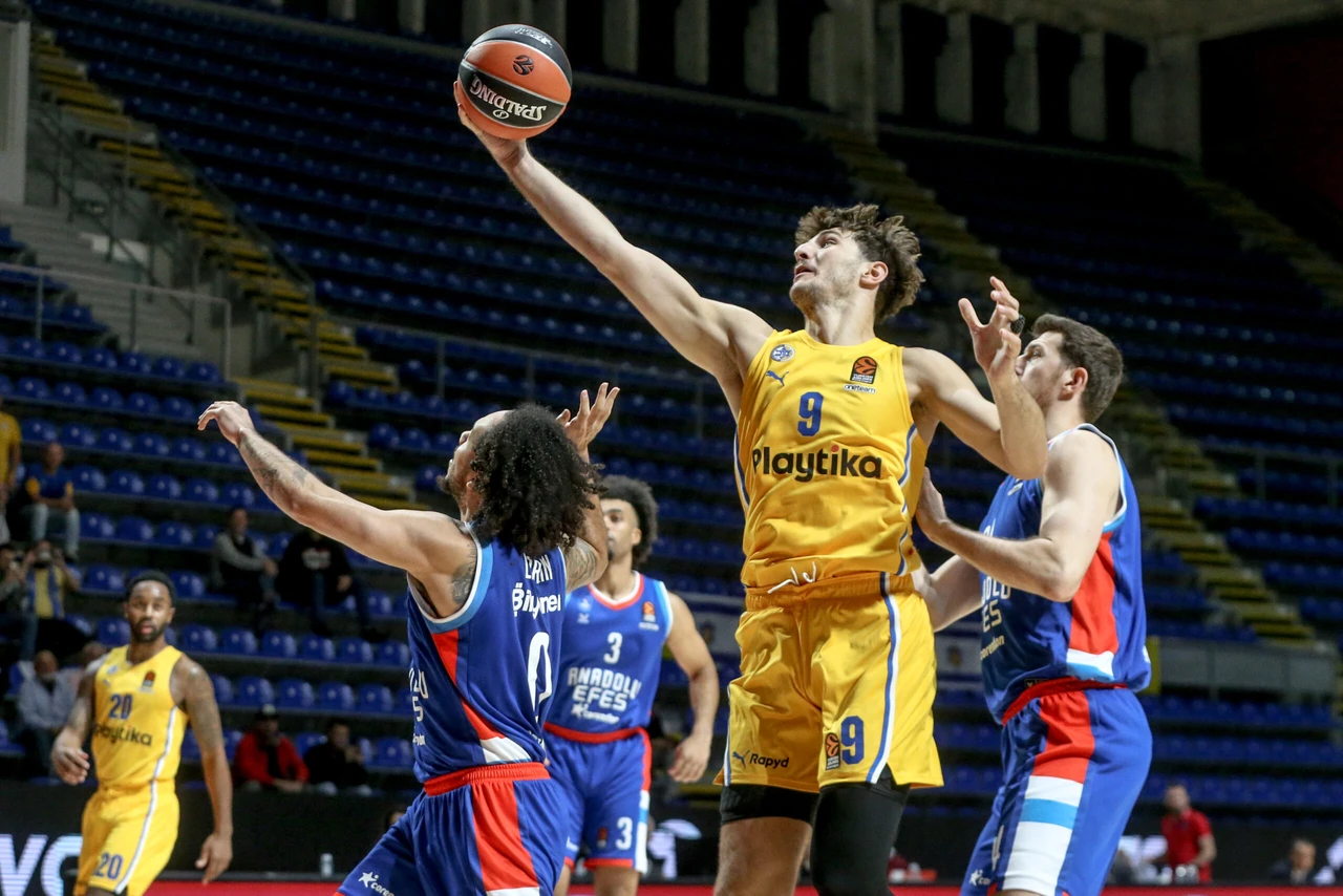 Maccabi Playtika player Roman Sorkin (#9) in yellow uniform reaches for the ball against Anadolu Efes defenders during Week 3 of the Turkish Airlines EuroLeague match at Aleksandar Nikolic Hall in Belgrade, Serbia. The game was played on neutral ground, with Sorkin elevating above blue-uniformed Anadolu Efes players attempting to block his shot