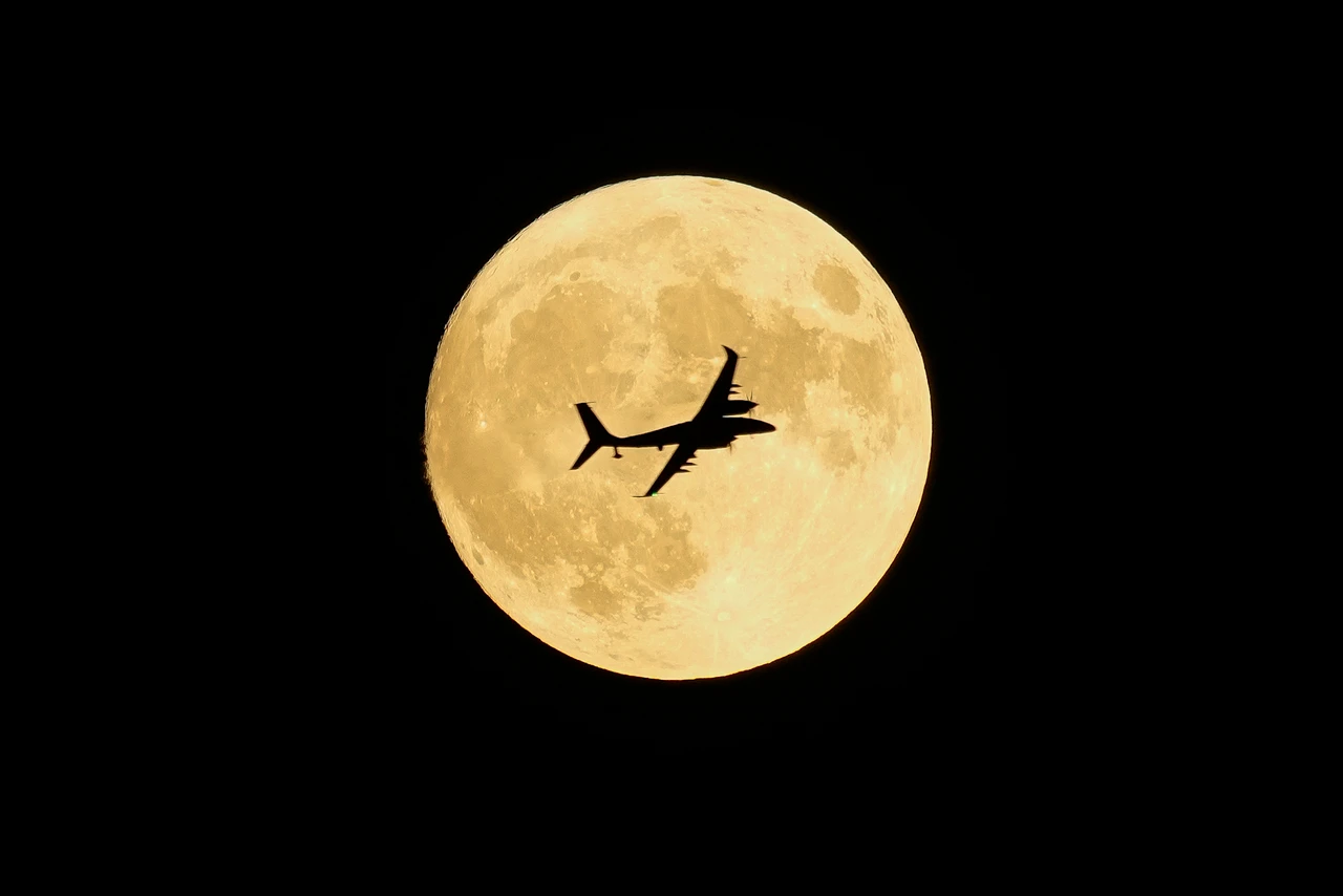 The silhouette of a Bayraktar AKINCI unmanned combat aerial vehicle (UCAV) flying across a full moon against a black night sky. The drone's distinctive shape with its long wingspan and twin tail booms is clearly visible as a dark outline against the bright, golden-yellow surface of the moon