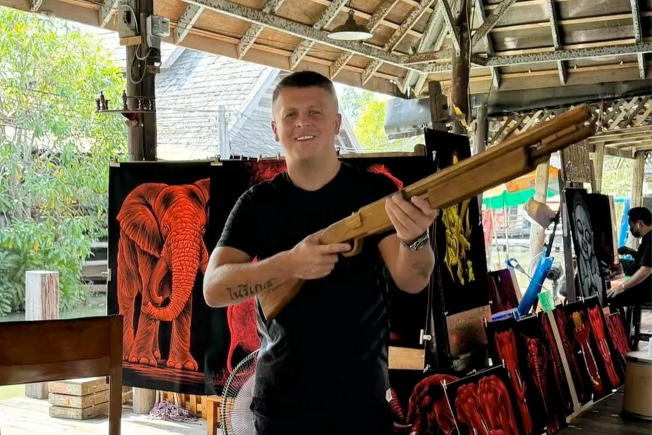 Ewan Corbett, dressed in a black T-shirt, holds a wooden rifle and smiles while standing in a market with red elephant-themed artwork in the background.