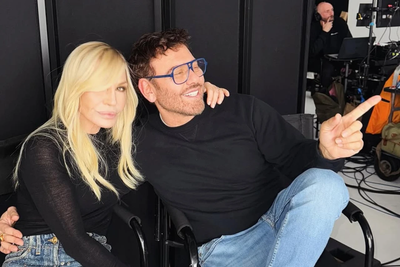 Donatella Versace sits beside photographer Mert Alas, both dressed in black, as they smile and pose for the camera in a relaxed studio setting.