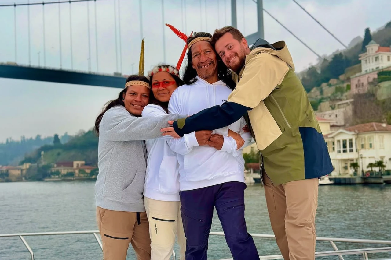 Three Waorani visitors wearing white hoodies and traditional feathered headbands stand with Alper Rende in front of the Bosphorus Bridge, embracing each other.