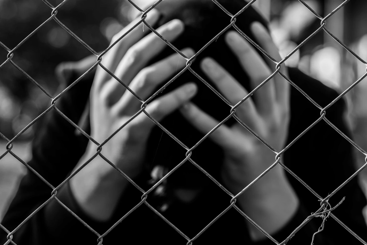 A distressed teenage boy holds his head behind a wired fence at a correctional facility in a black-and-white conceptual image highlighting juvenile delinquency.