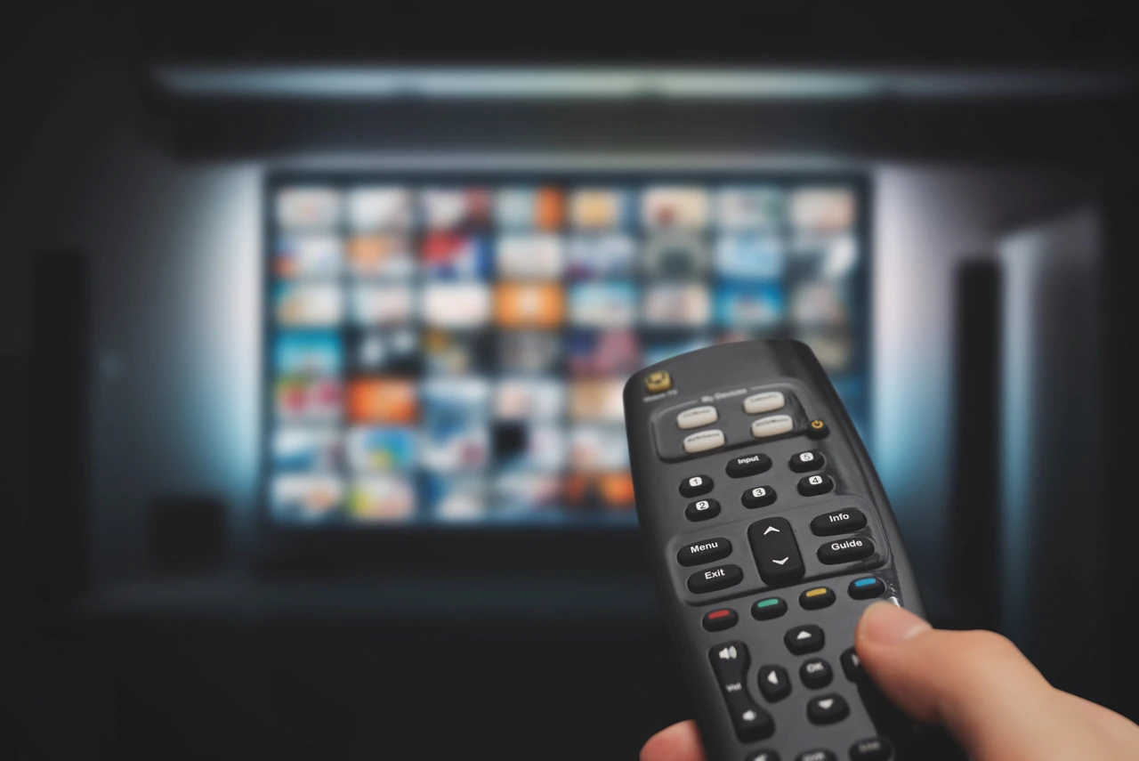 A hand holding a black remote control pointed toward a television screen in the background. The TV displays a blurred grid of colorful thumbnails representing various streaming content options. The image is taken in a dimly lit room with the screen providing most of the illumination