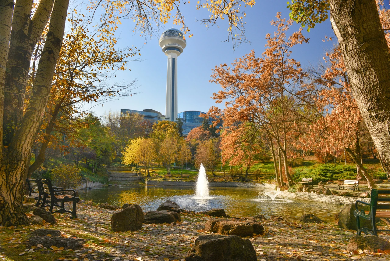 Photo shows Botanic Park in Ankara during autumn, featuring a pond with fountain surrounded by trees with yellow and orange foliage, with Atakule Tower visible in the background against a clear blue sky