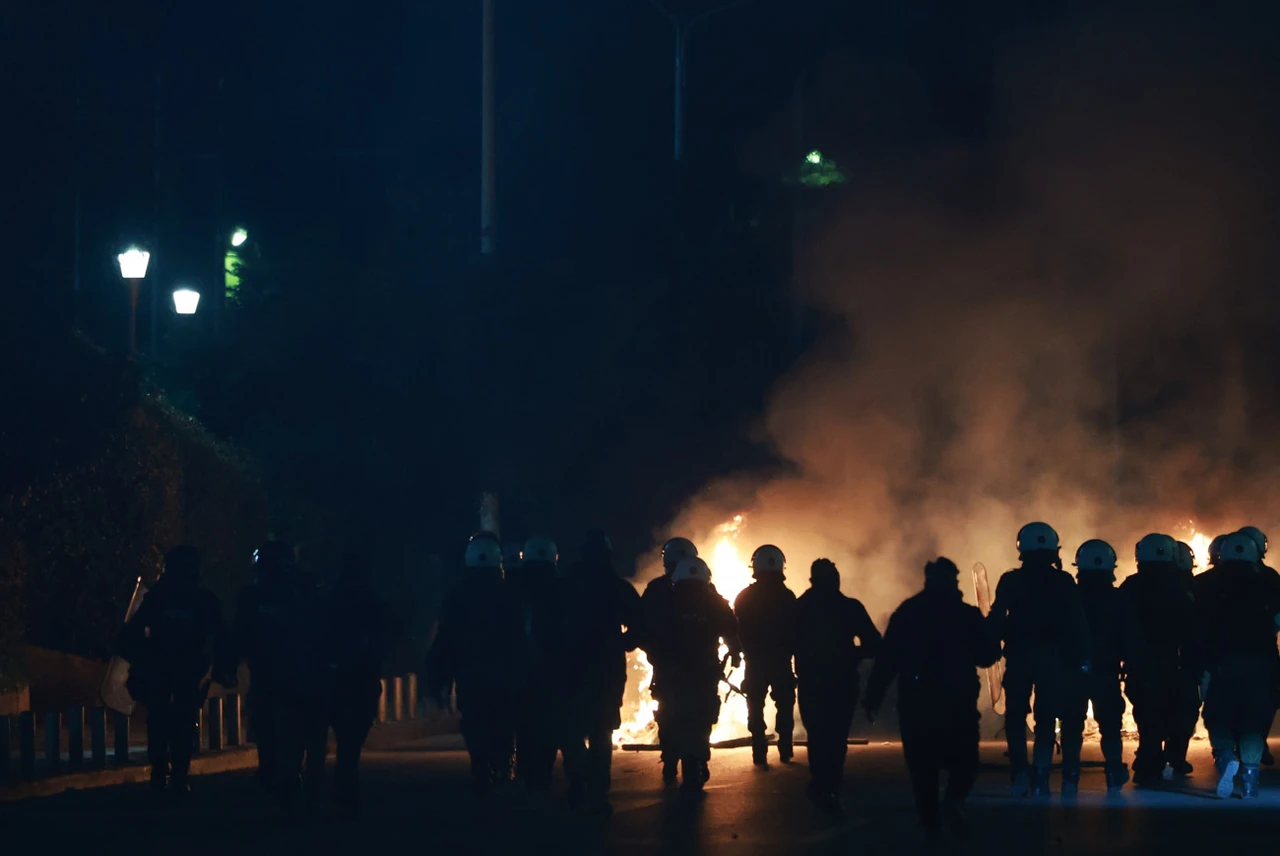 Riot police officers in helmets and protective gear walk towards a fire in the middle of the road during protests in Athens over the Tempi train accident.