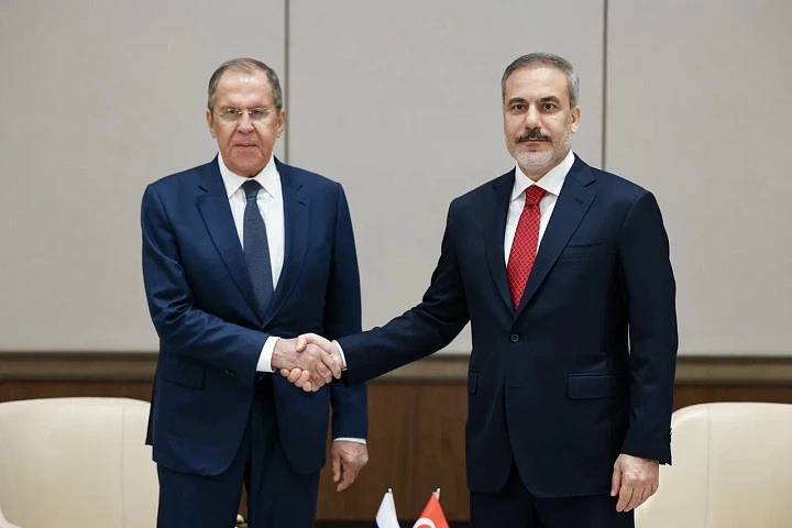 Photo shows Russian Foreign Minister Sergey Lavrov (left) and Turkish Foreign Minister Hakan Fidan in dark business suits shake hands against a beige background. On the left is an older man with gray hair wearing a navy suit and blue tie. On the right is a younger man with a mustache wearing a black suit and red tie. Small national flags can be seen in the background