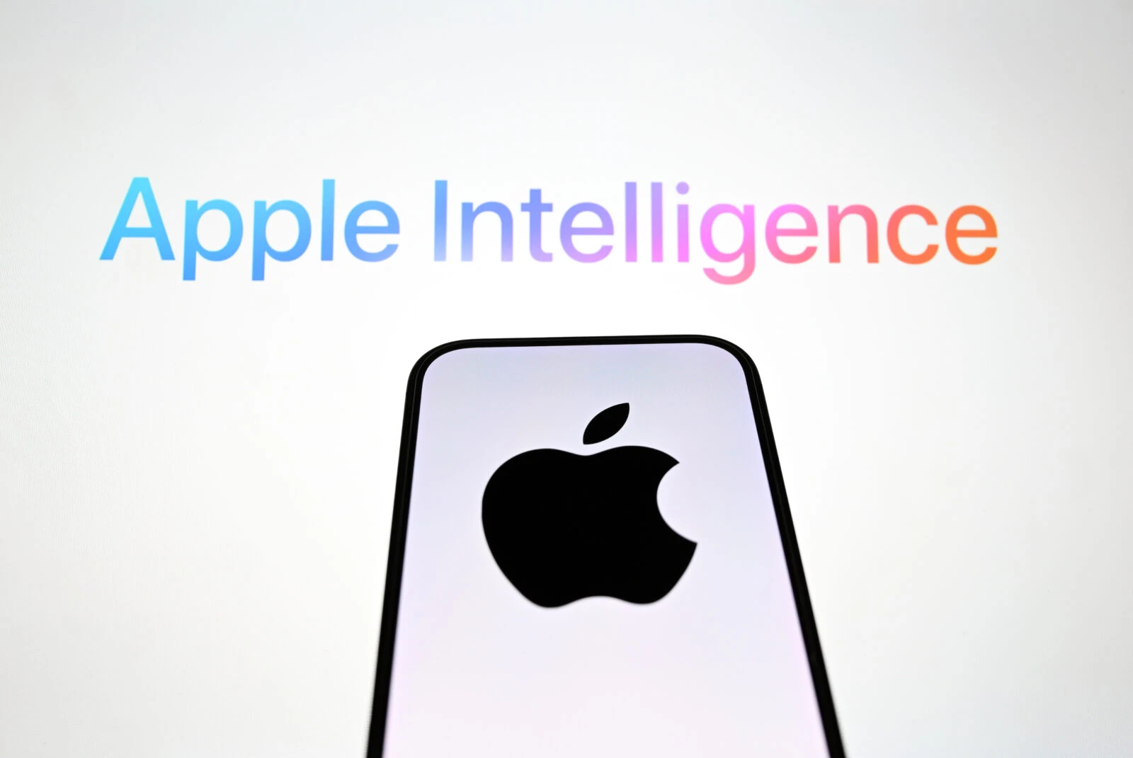 In this photo illustration, the 'Apple' logo is displayed on a mobile phone screen in front of a computer screen displaying Apple Intelligence logo