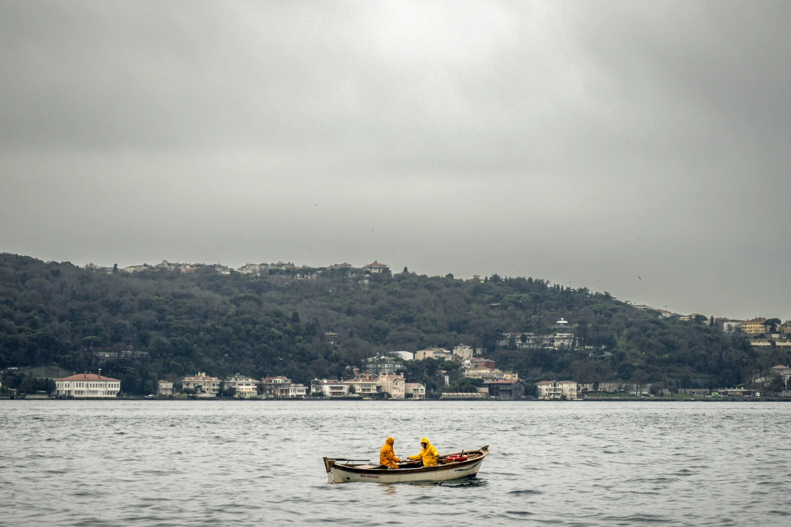 Two fishermen in bright yellow raincoats row a small wooden boat across the waters of Turkey's Bosphorus