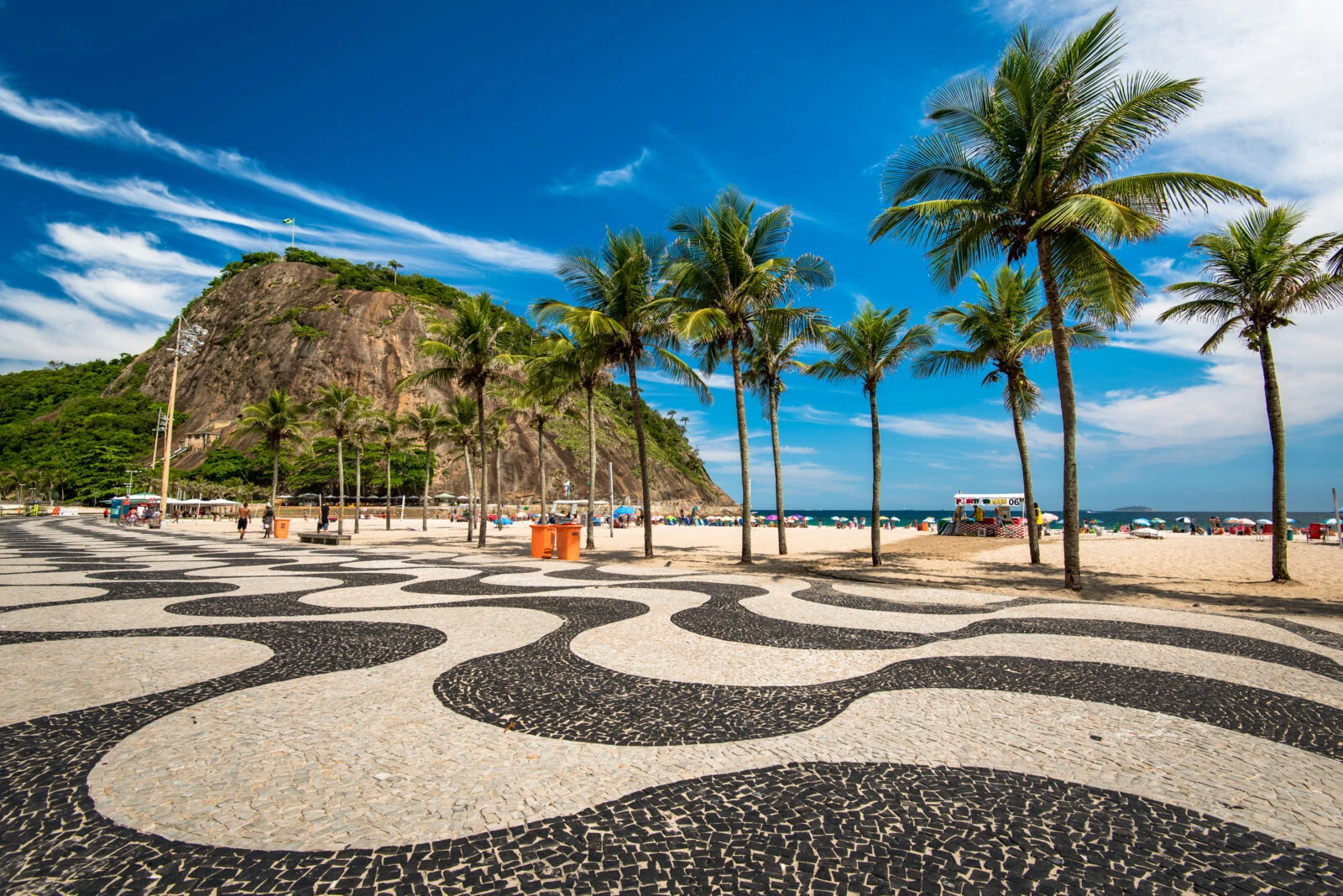 A detailed shot of Copacabana Beach’s wave-patterned pavement with palm trees, mountains, and the Atlantic Ocean in the background