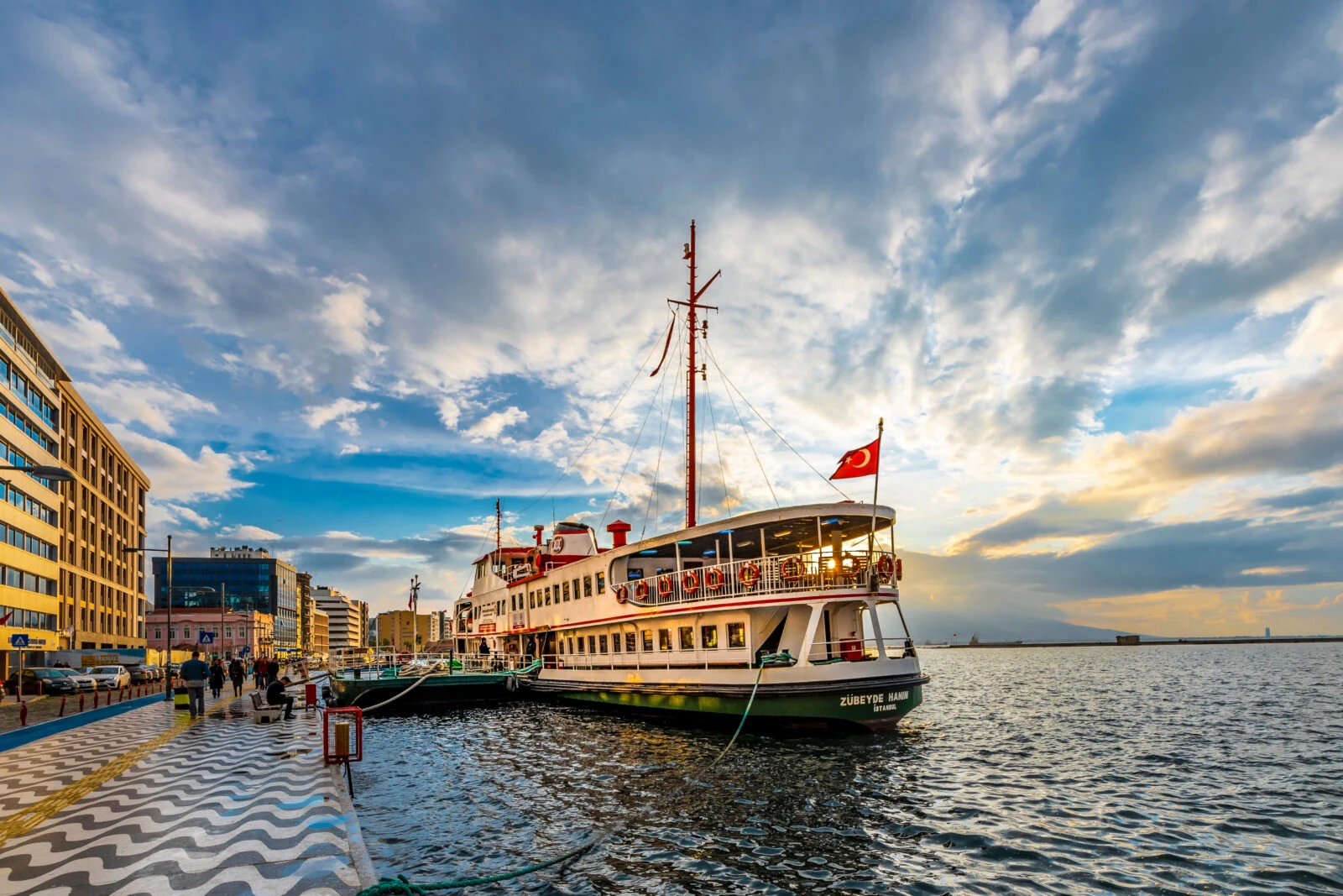 A ferry with the Turkish flag docked at Izmir’s Kordon, with the wave-patterned promenade, cityscape, and a dramatic sunset sky in the background