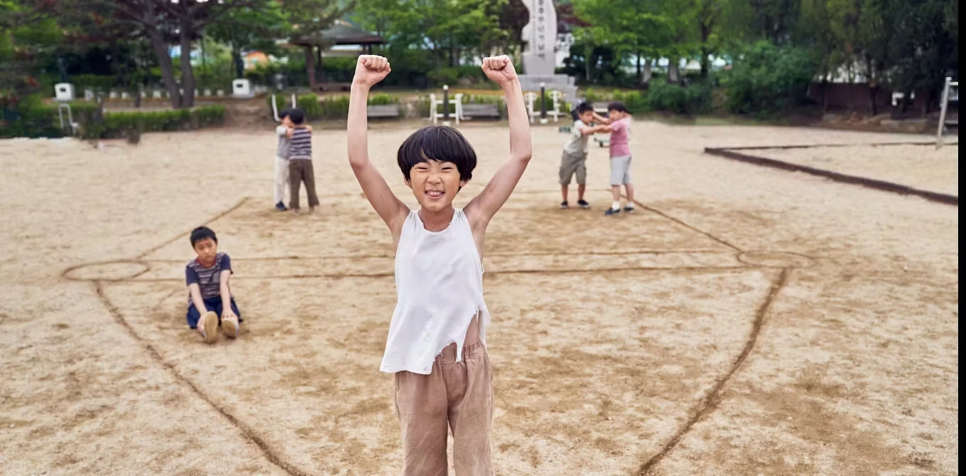 A group of children playing squid-like games in an outdoor park, with one child raising their arms in celebration in the foreground