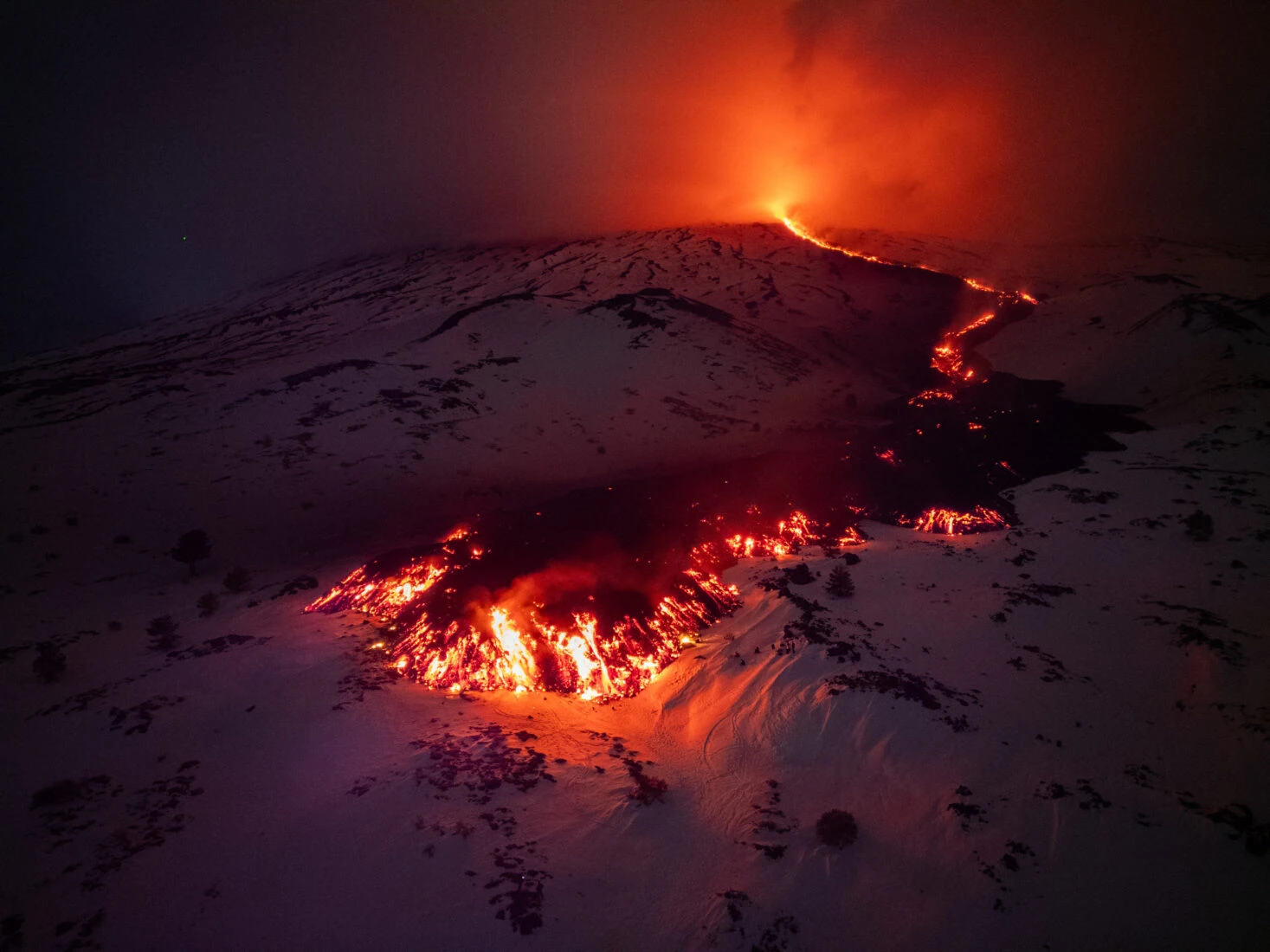 A vivid stream of molten lava flows from a fissure on Mount Etna during an eruption at night, casting a fiery glow against the dark sky.