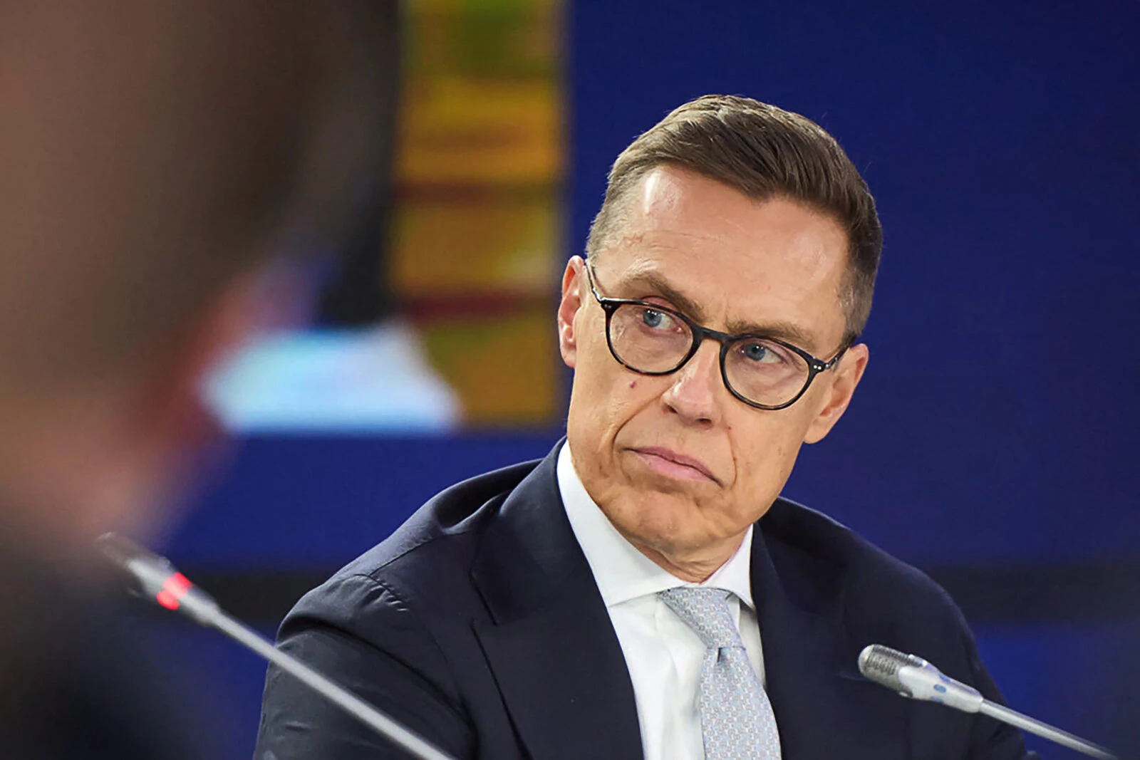 A photograph of Finland's President Alexander Stubb shows him in a formal setting, wearing a navy-blue suit, white dress shirt, and a light-colored tie. He has short, neatly combed brown hair and wears black-rimmed glasses. His expression is serious and focused as he listens intently to someone speaking. A microphone is visible in front of him, and the blurred background suggests an international or diplomatic event.