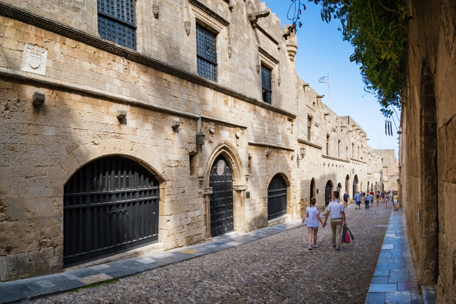 A couple walks hand in hand down the medieval Street of the Knights (Ippoton), surrounded by centuries-old stone buildings in the Old Town of Rhodes, Greece.