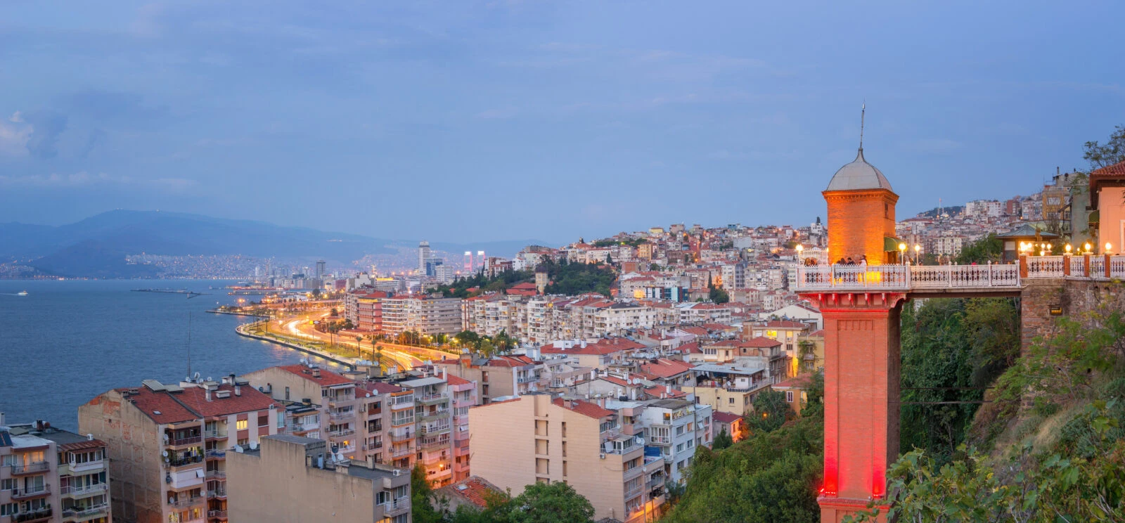 ramadan in izmir: A panoramic view of Izmir, Türkiye, at dusk, featuring the historic Asansör (Elevator) with its red-lit tower and viewing platform, overlooking a cityscape with coastal roads, residential buildings, and the Aegean Sea in the background.