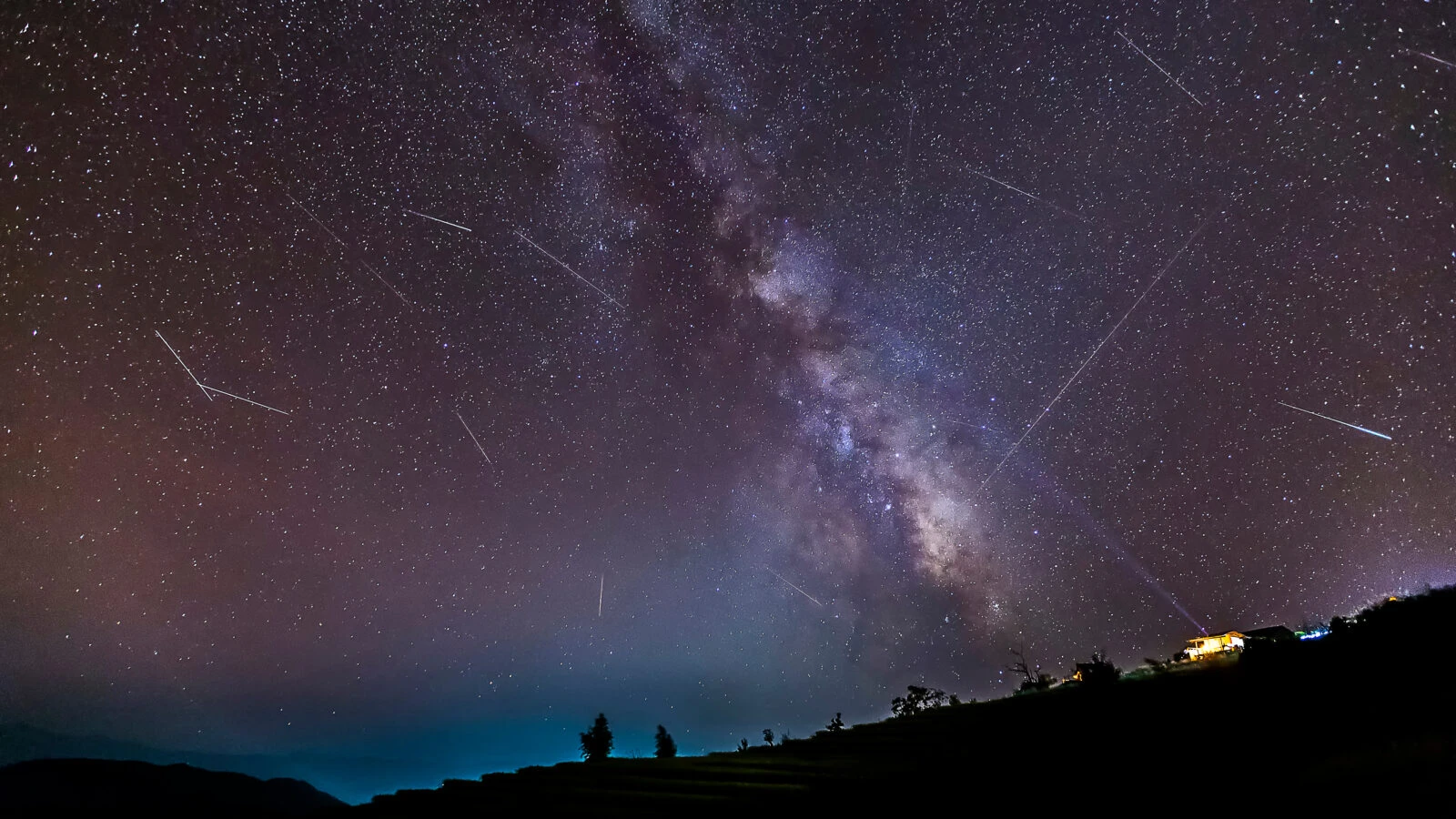 A dazzling night sky filled with streaking meteors above a mountainous landscape.