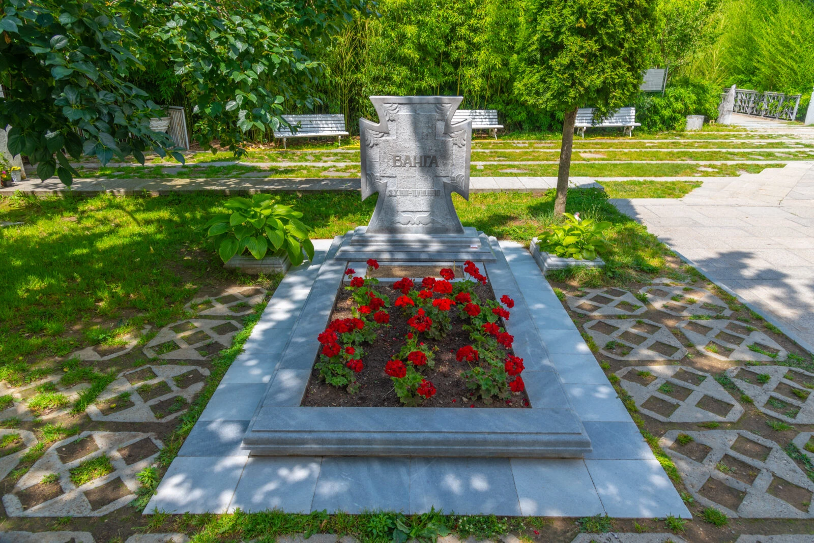 The grave of Baba Vanga, marked by a stone cross-shaped headstone inscribed with her name, surrounded by red flowers and set in a tranquil garden with benches and greenery.