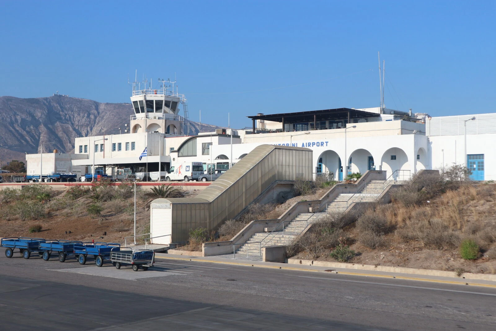 A wide-angle view of Santorini International Airport taken from the apron, showcasing the terminal building and surrounding infrastructure under a clear sky.