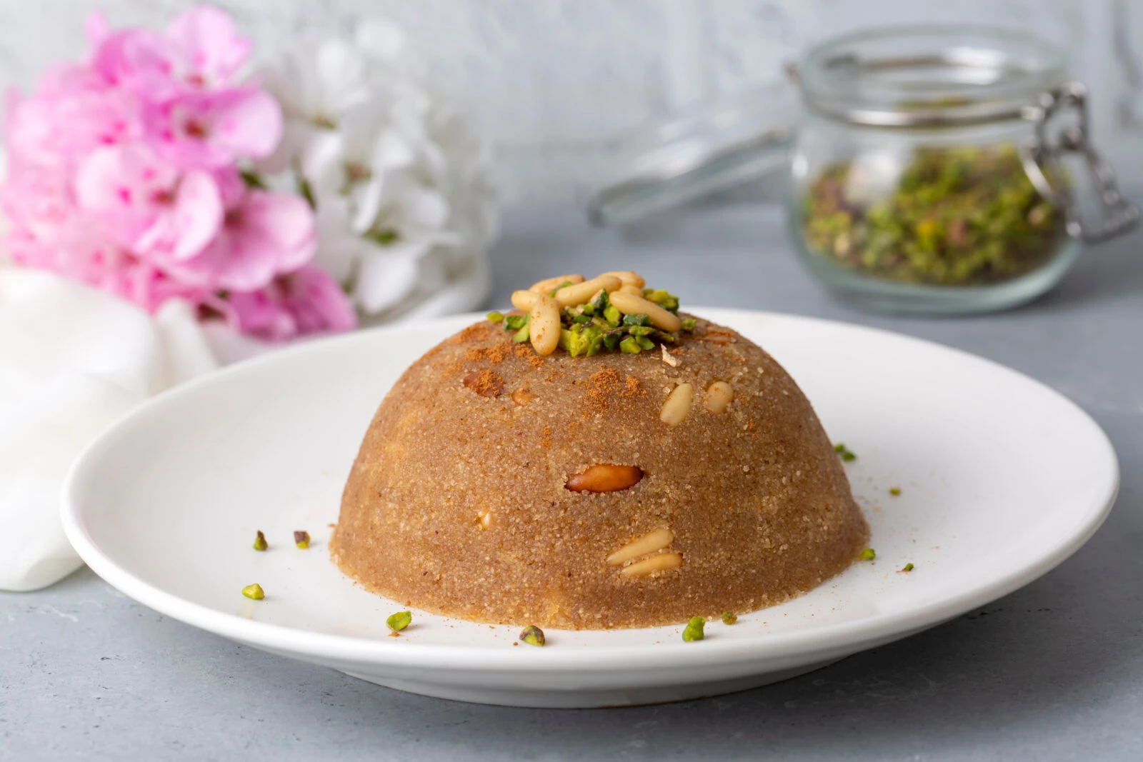 A plate of Irmik Helvasi, a Turkish semolina dessert garnished with pine nuts and pistachios, set on a table with flowers in the background.