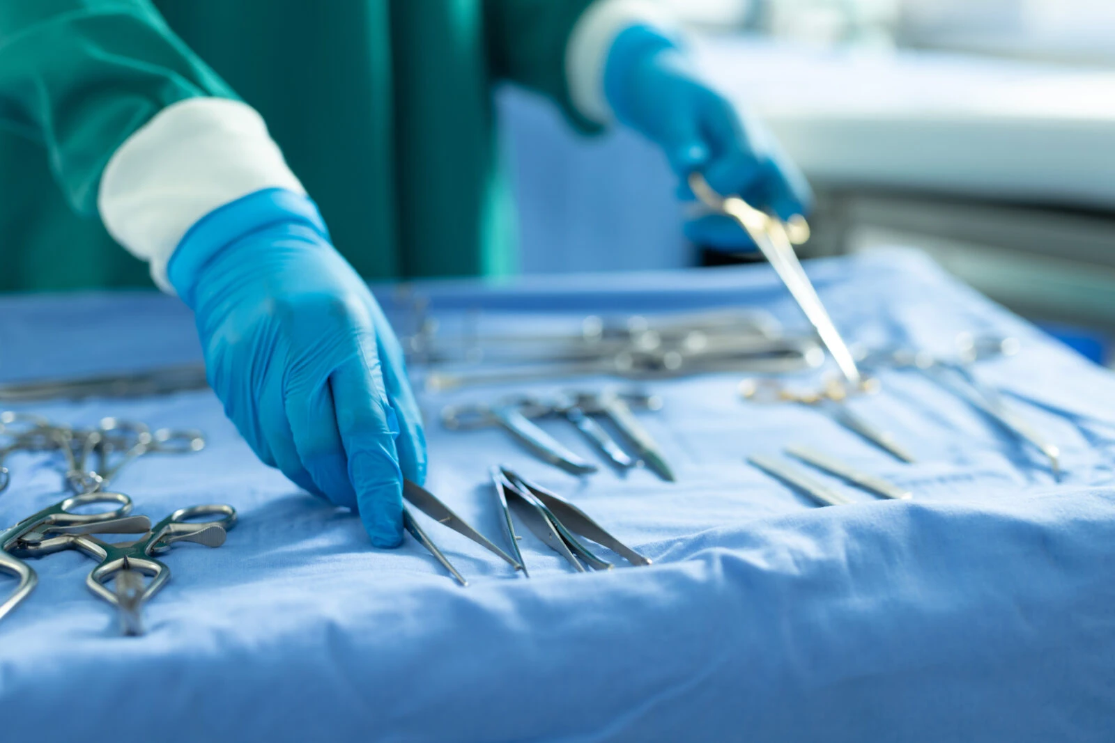 A set of surgical tools laid out on a sterile blue table in an operating room, with blurred surgeons in blue scrubs performing a procedure in the background.