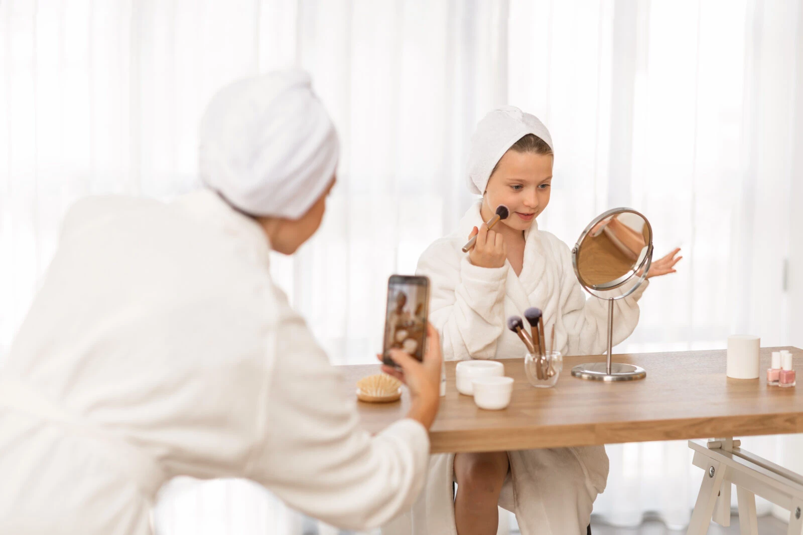 A mother and her young daughter in bathrobes record a makeup tutorial at home. The daughter applies makeup while the mother films the process with a smartphone. - sharenting