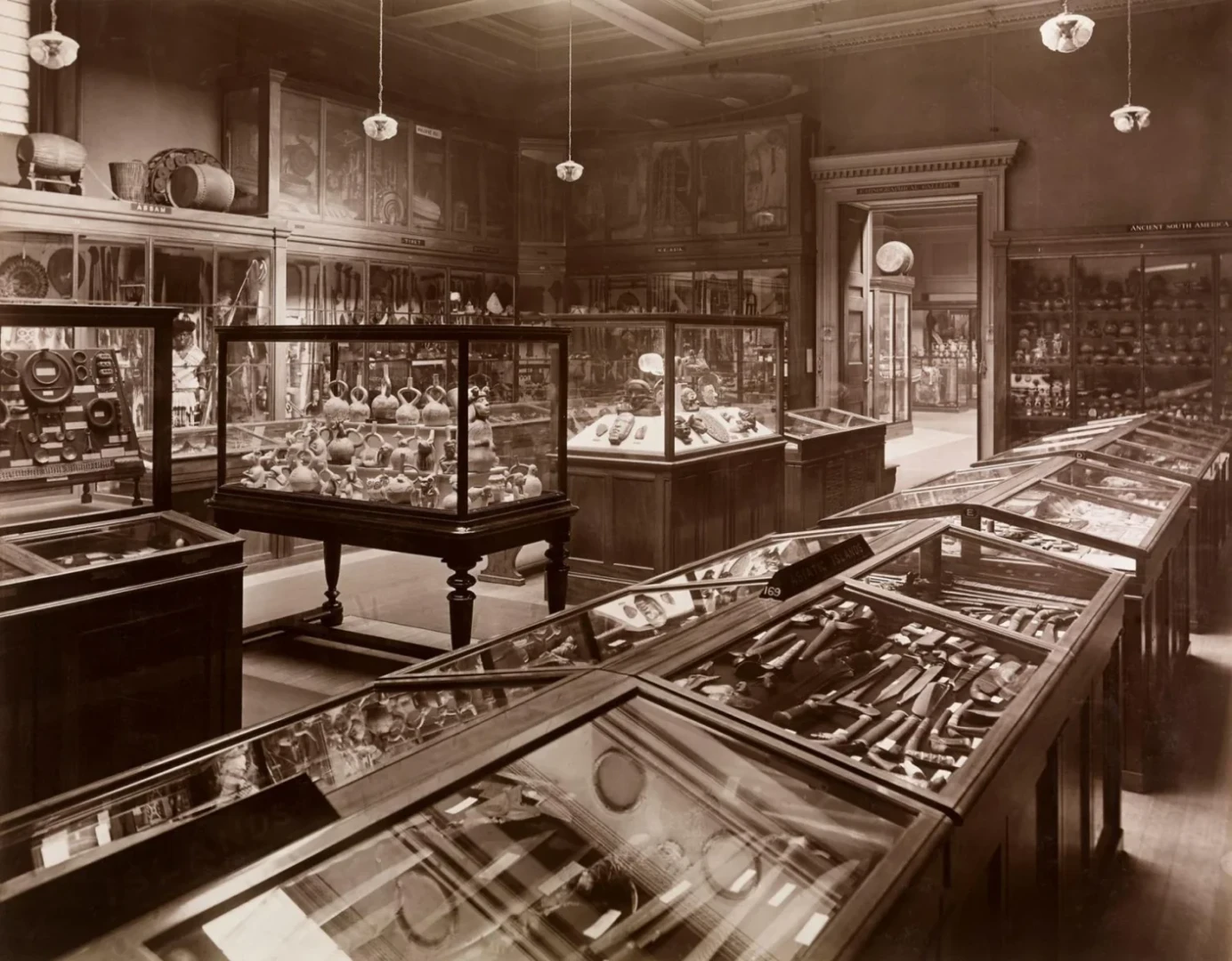 A historical photograph of the Ethnographic Gallery at the British Museum, taken around 1923. The image showcases display cases filled with artifacts and exhibits from various cultures, arranged in a grand exhibition hall.