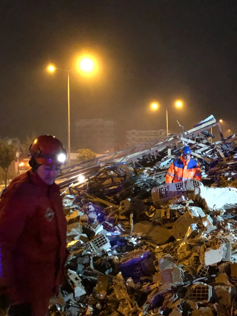 Rescue workers in red and orange gear search through debris at night, illuminated by streetlights in turkey