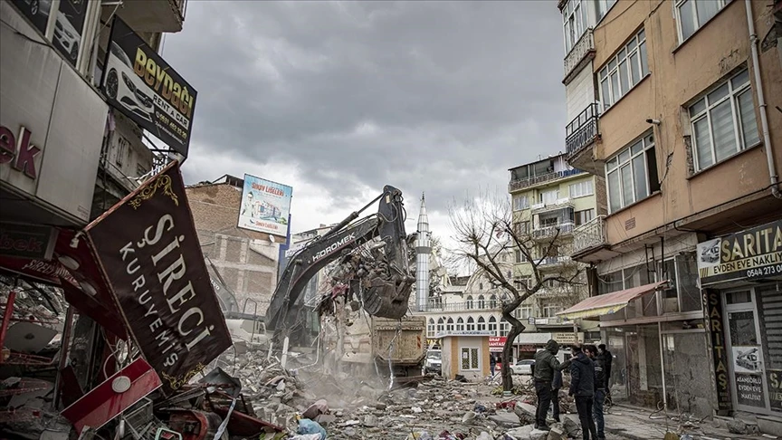 A backhoe is clearing debris as people stand nearby, observing the aftermath of the disaster