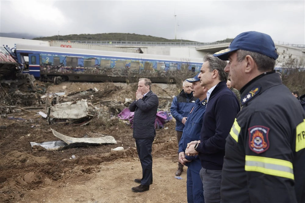 Greek Prime Minister Kyriakos Mitsotakis and Minister of Infrastructure and Transport Kostas Karamanlis stand at the site of the train crash in Tempi, Greece, alongside emergency officials. The wreckage of a derailed train and debris from the accident are visible in the background. Karamanlis resigned later that day