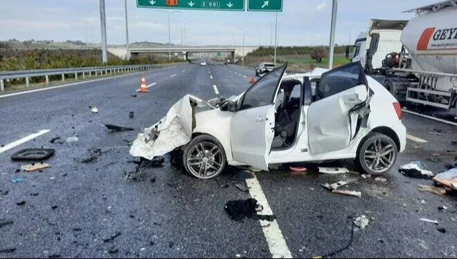 A severely damaged white car on a highway with debris scattered around, emergency cones placed in the background, and a tanker visible on the side.

