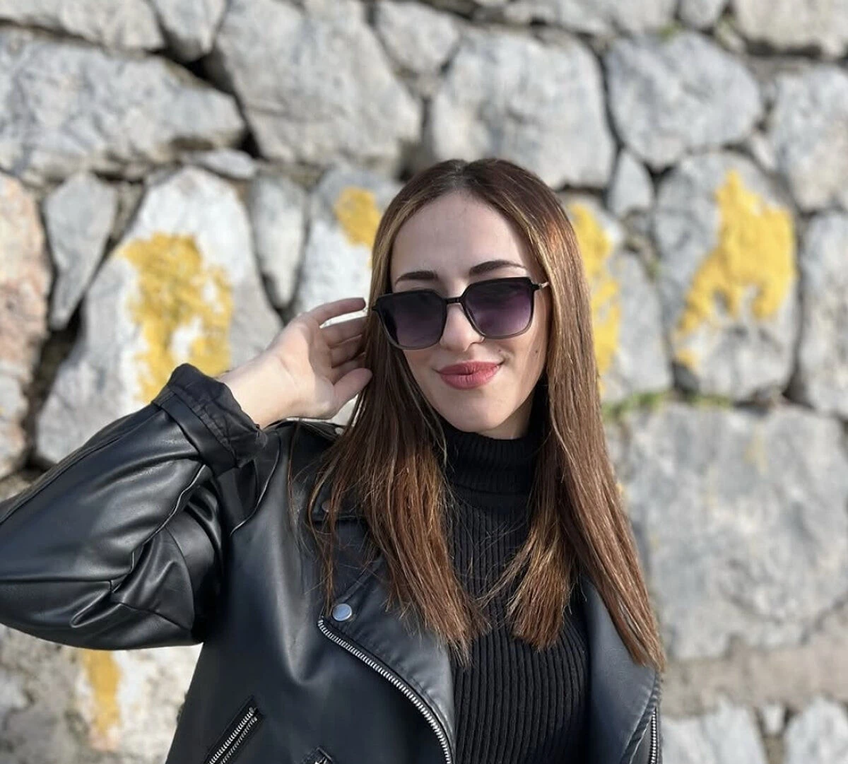 A young woman with long brown hair wearing a black leather jacket and sunglasses, smiling as she poses in front of a stone wall.