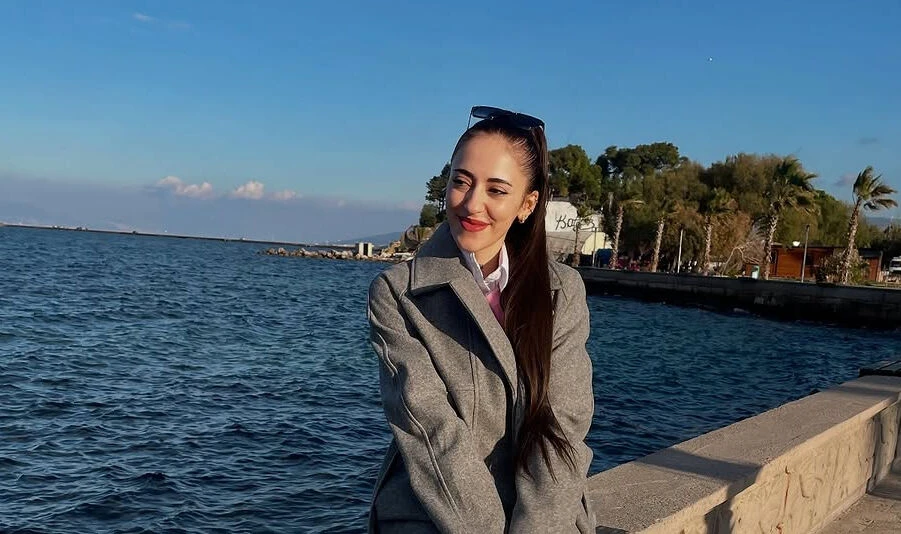 A young woman in a gray coat standing by the waterfront, smiling with the sea, palm trees, and a clear blue sky in the background.