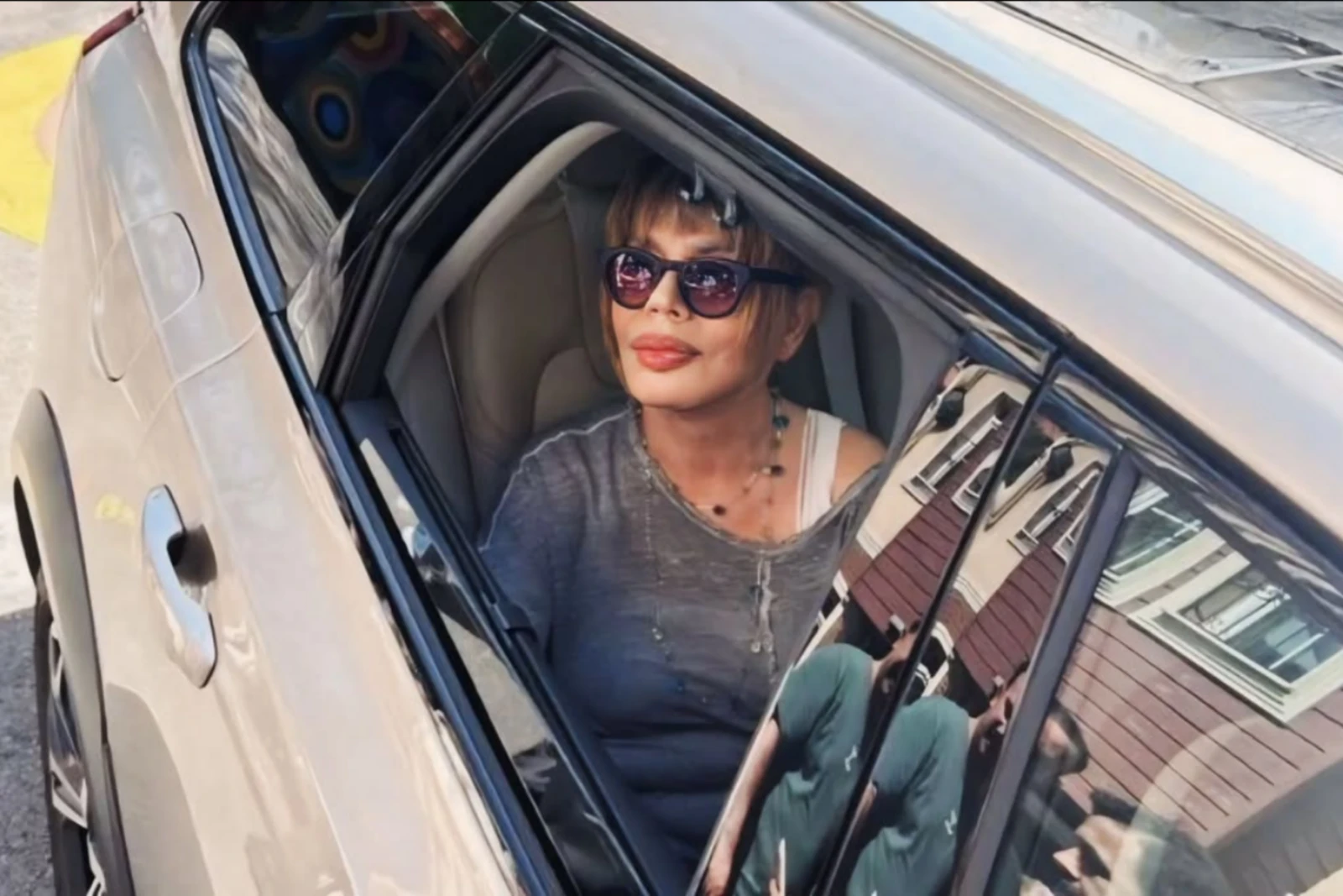 Sezen Aksu looking out from a car window, wearing sunglasses and a gray top with a necklace.