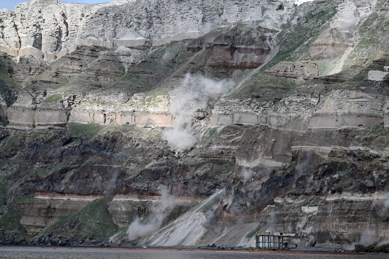 A massive cloud of dust rises over a rocky cliff on the island of Santorini after a landslide, caused by ongoing seismic activity. The rugged coastline and turbulent sea reflect the impact of the tremors in the Aegean region.