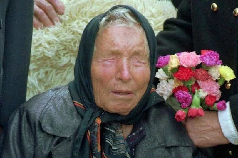 An elderly Baba Vanga wearing a black headscarf and dark clothing, holding a bouquet of colorful flowers, with her eyes closed as she is surrounded by people.