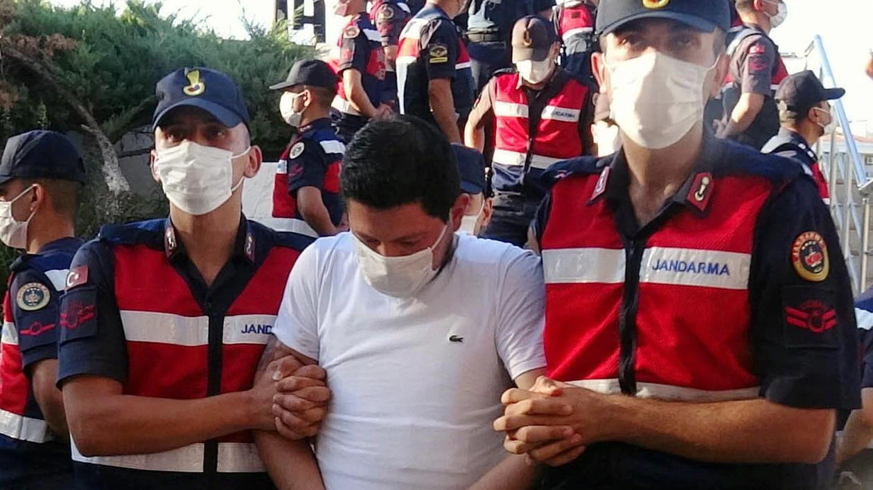 killer Cemal Metin Avci, wearing a white t-shirt and face mask, being escorted in handcuffs by two uniformed gendarmerie officers