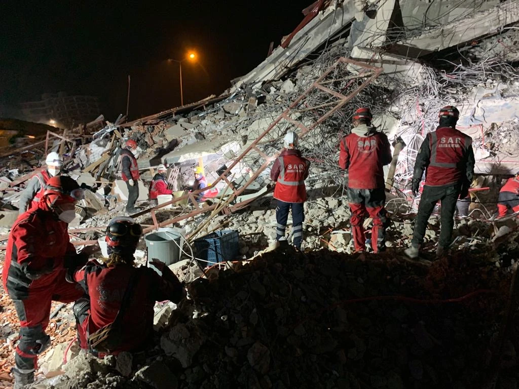 A group of search and rescue workers in red uniforms and protective gear carefully navigate the rubble of a collapsed building in Antakya, Türkiye