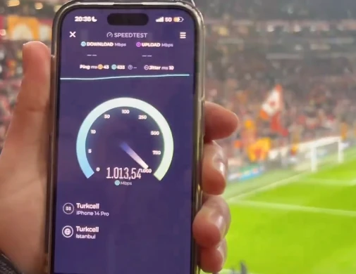 A person holds an iPhone 14 Pro inside a stadium, displaying a speed test result on the screen