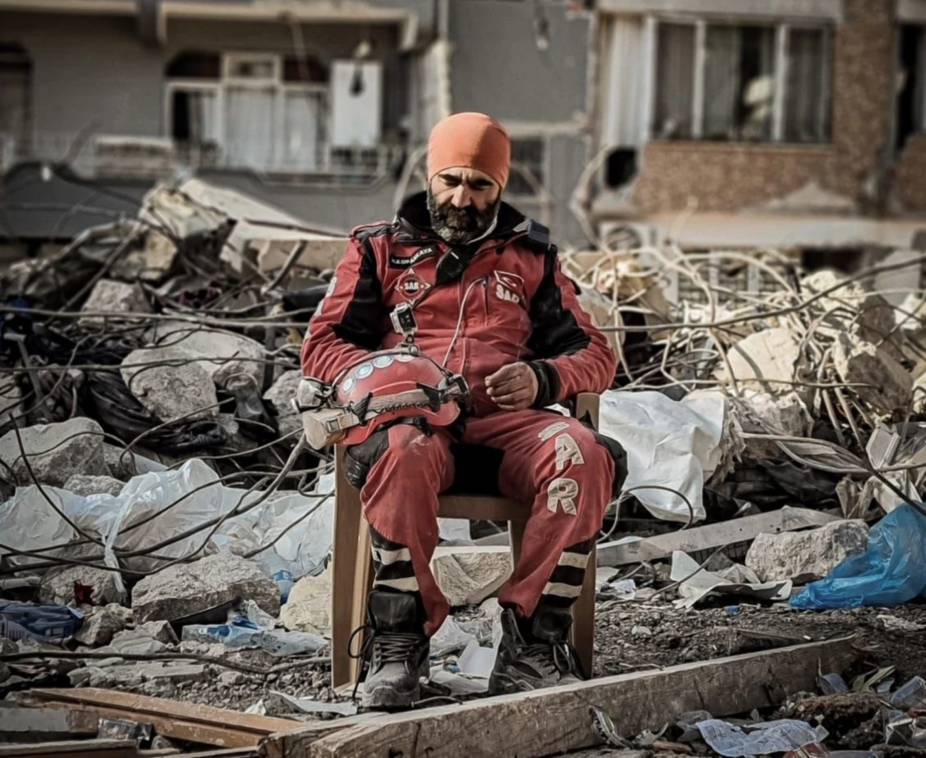 A SAR rescue worker in a red uniform and orange beanie sits on a chair in the middle of debris, holding his helmet, with collapsed buildings in the background