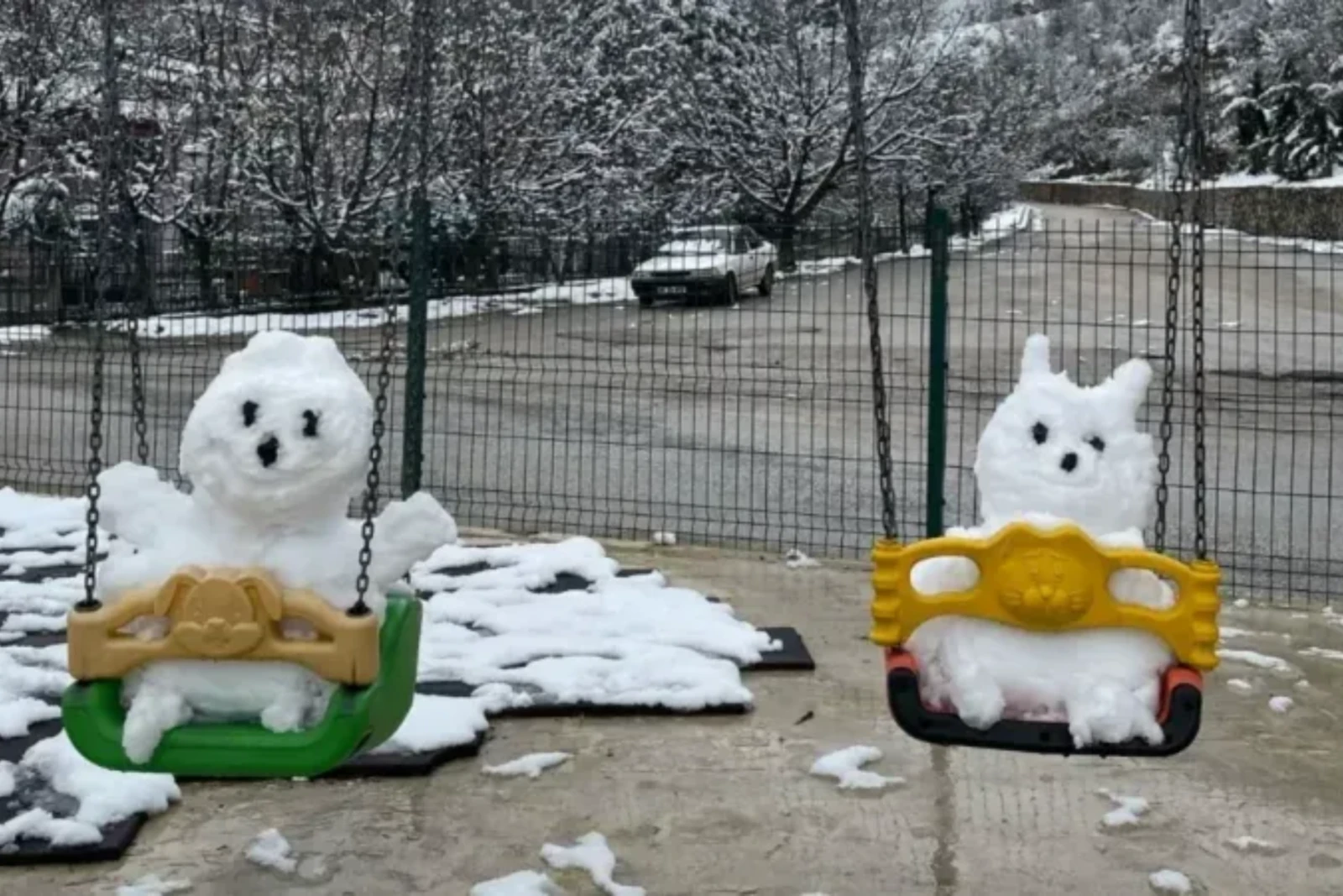 Two small snowmen, one resembling a rabbit and the other with a ghostly face, are placed on swings in a playground covered in snow.